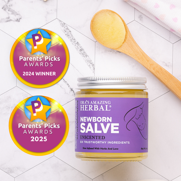 A jar of ORA's Amazing Herbal Newborn Salve with a purple label flat lay on a white marble surface next to a wooden spoon holding yellow salve. Two circular Parents' Picks Awards 2024 Winner and 2025 badge graphics are shown on the left.