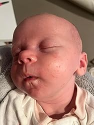 Newborn baby with a bald head and redness in face wearing light-colored outfit.