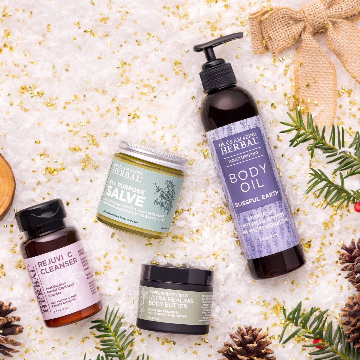 Four skincare products from Blush Garden Herbal are arranged in a flat lay on a white textured surface with gold foil flakes, evergreen branches, pinecones, and a burlap ribbon. The products include Rejuvenating Cleanser, Almond Rose Salve, Deep Healing Ultra-Soothing Body Butter, and Bluefulse Earth Body Oil.