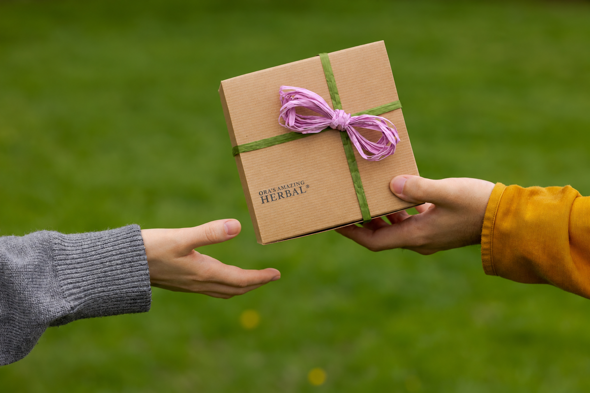A person in a gray sweater extends their hand to receive a small brown kraft gift box tied with a green ribbon and a purple bow, held by another person wearing a yellow sleeve, against a blurred green grass background.