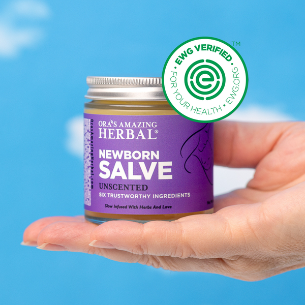 A hand holding Ora's Amazing Herbal Newborn Salve against a bright blue sky background with white clouds. A green circular EWG Verified badge is overlaid on the top right of the jar.