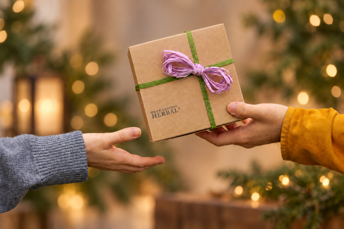 A person in a gray sweater extends a hand while another person in a mustard yellow sleeve holds a small brown kraft gift box tied with a purple ribbon. The background is a softly blurred holiday scene with warm lights and greenery.