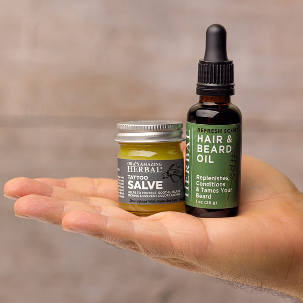 Hand holding a jar of herbal tattoo salve and a bottle of hair and beard oil against a blurred background