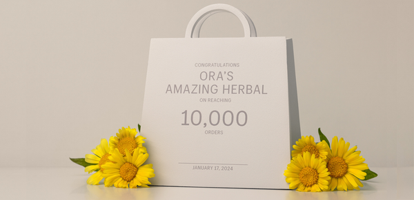 A white paper shopping bag plaque with printed text celebrating 10,000 orders, styled with yellow daisy flowers on a light beige background.