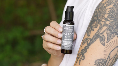 A person holding a bottle of Herbal Tattoo Oil Unscented against a blurred green outdoor background. The person has a black floral tattoo on their upper arm and is wearing a white sleeveless top.
