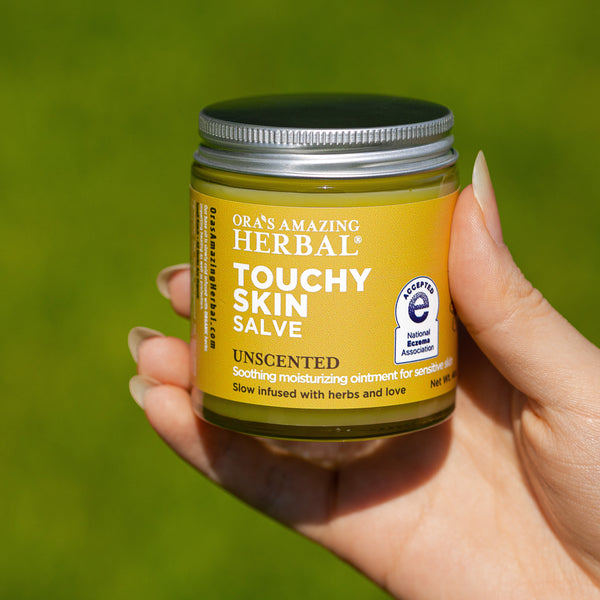 Hand holding a jar of Ora's Amazing Herbal Touchy Skin Salve against a green background
